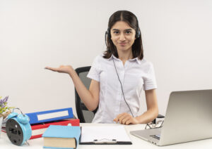 young-girl-white-shirt-headphones-presenting-something-with-arm-her-hand-smiling-sitting-table-with-folders-laptop-white-wall